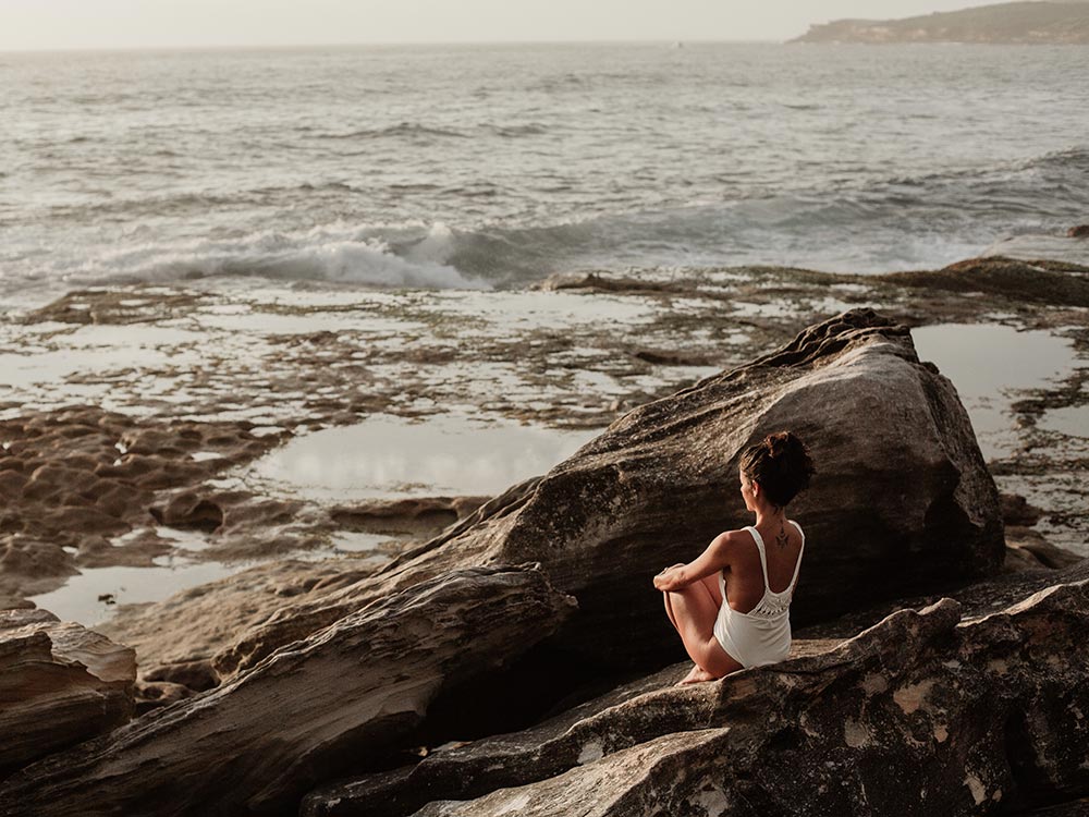 Femme en maillot blanc assise sur des rochers, contemplant l'océan au lever du soleil.