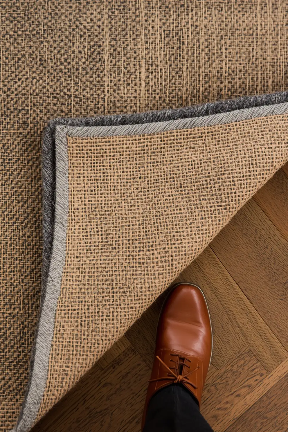 Tapis d'entrée tressé de couleur beige et gris, angle retourné montrant le dessous, un homme porte un soulier marron élégant sur parquet chevron.