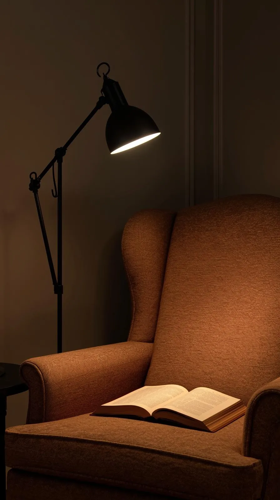 Un lampadaire noir orientable allumé, éclairant un livre ouvert posé sur un fauteuil en tissu dans l'obscurité.