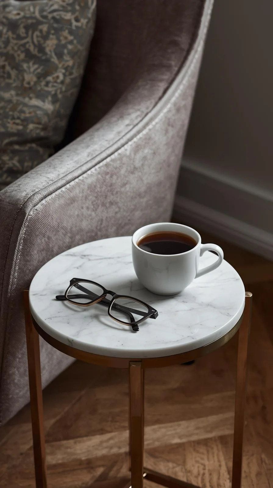 Une petite table d'appoint ronde avec un plateau en marbre et des pieds en laiton, portant une tasse de café et des lunettes.