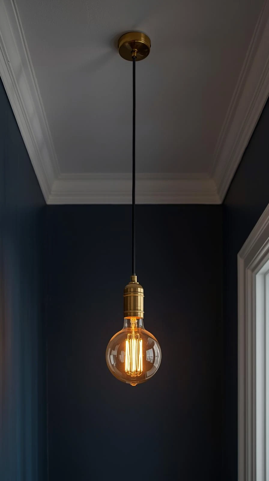 Une suspension luminaire fine avec un long câble noir, une douille en laiton et une ampoule Edison vintage, sur fond de mur bleu nuit.