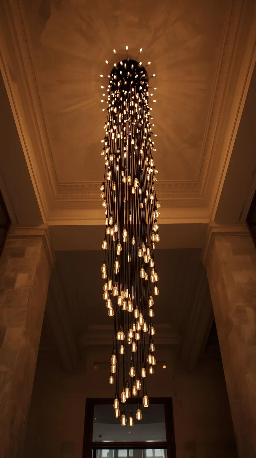 Un très grand lustre en spirale avec de multiples ampoules descendant sur plusieurs mètres dans un hall d'entrée à haut plafond.