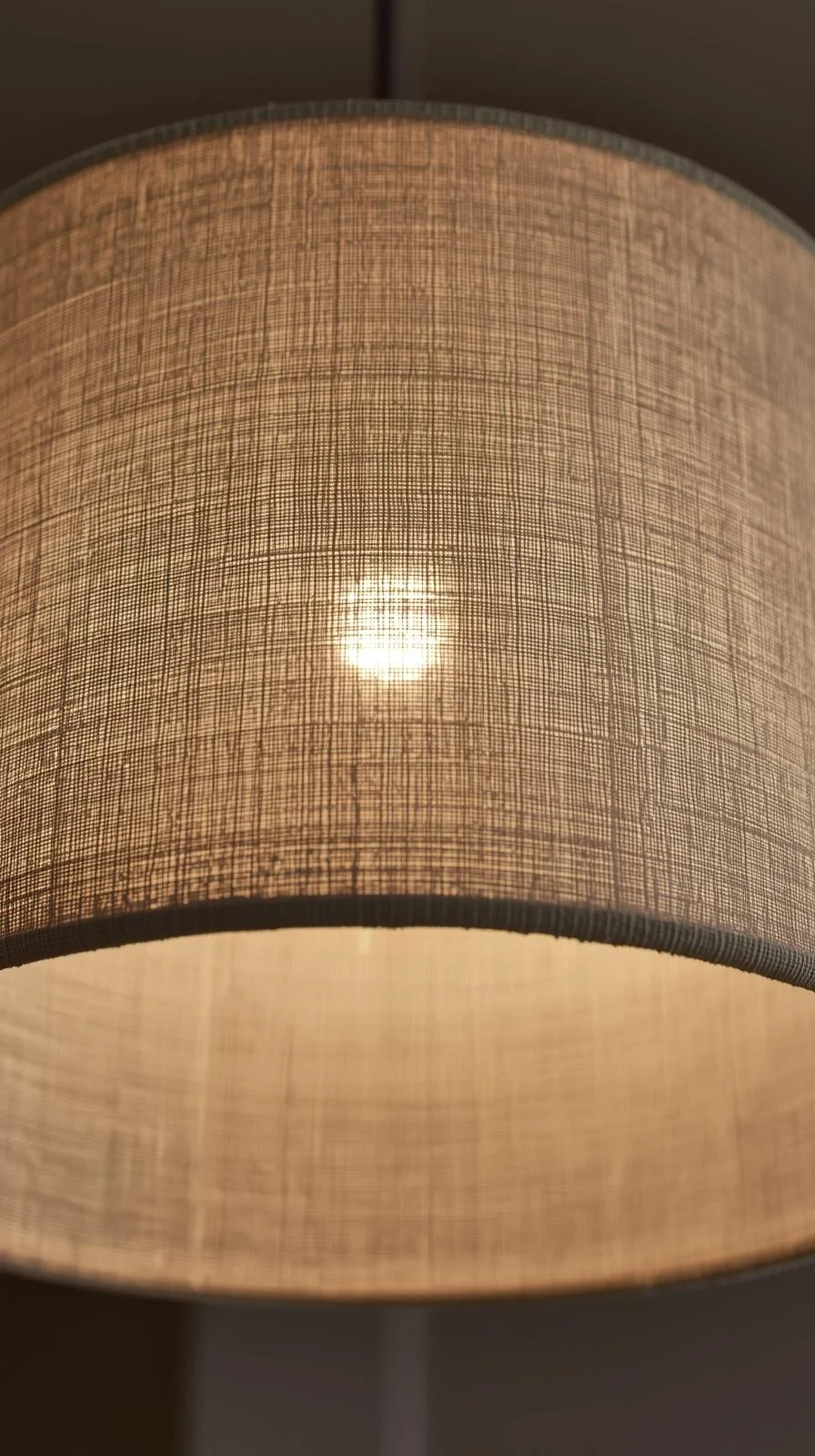 Gros plan d'un lustre tambour cylindrique recouvert de tissu en lin texturé (beige ou gris), allumé et diffusant une lumière douce.