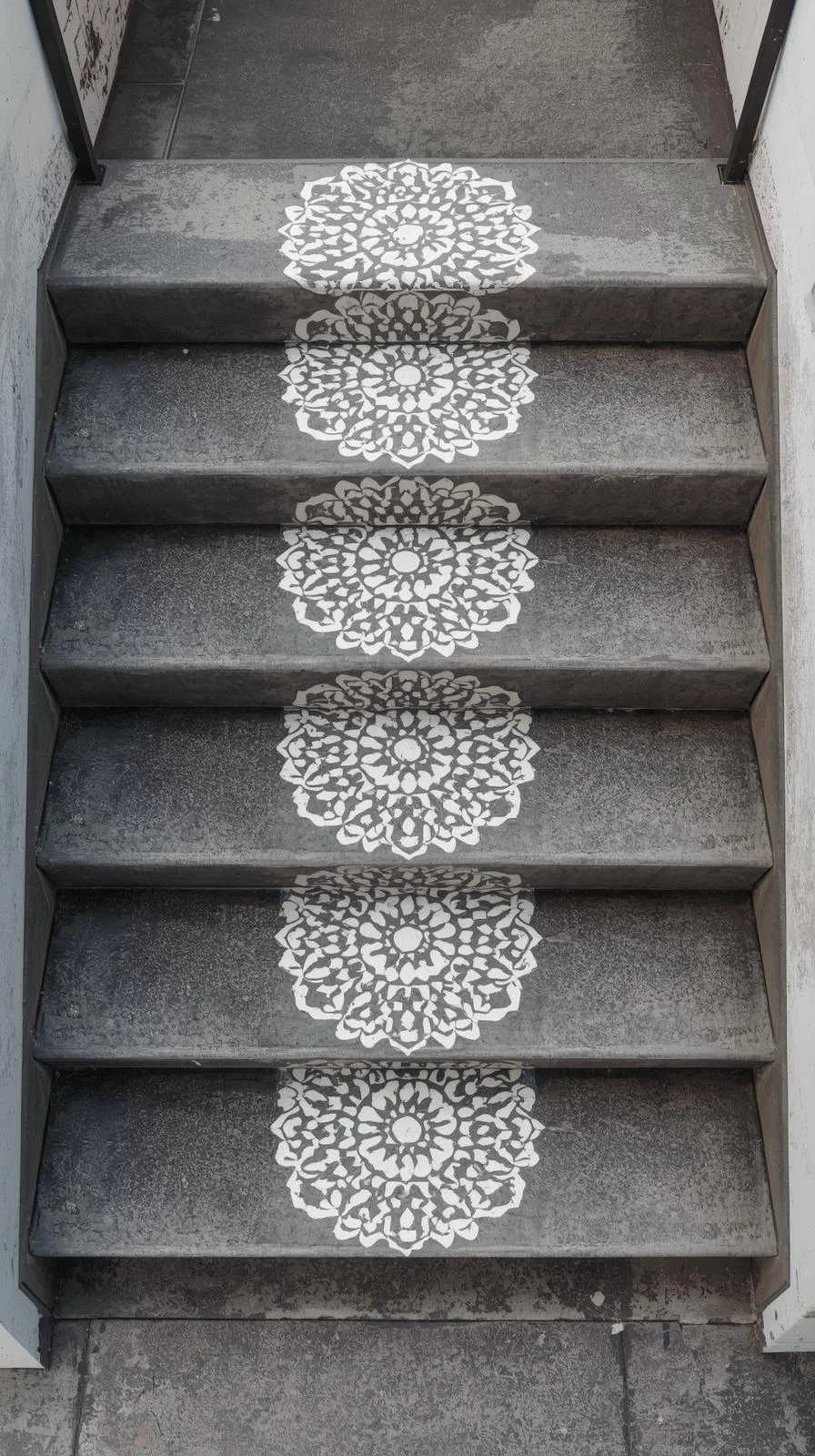 Vue plongeante d'un escalier en béton gris dont chaque marche est décorée au centre d'un grand motif de mandala blanc, peint au pochoir.