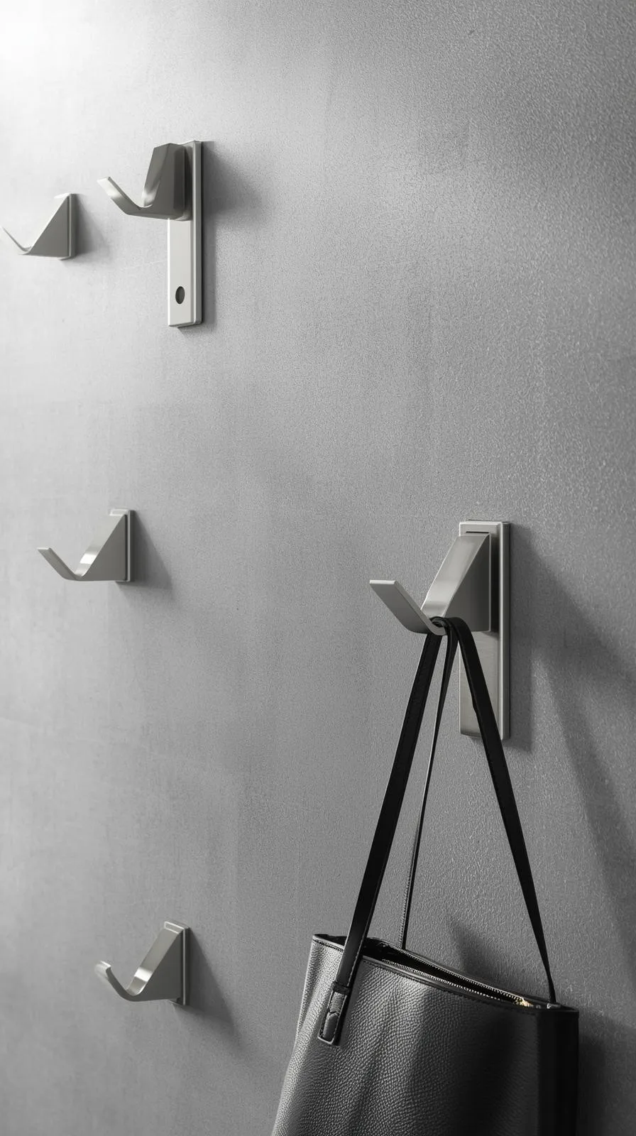 Gros plan sur des crochets pliables design en métal brossé, fixés sur un mur texturé gris. Un crochet est déplié et tient un sac, les autres sont repliés à plat.